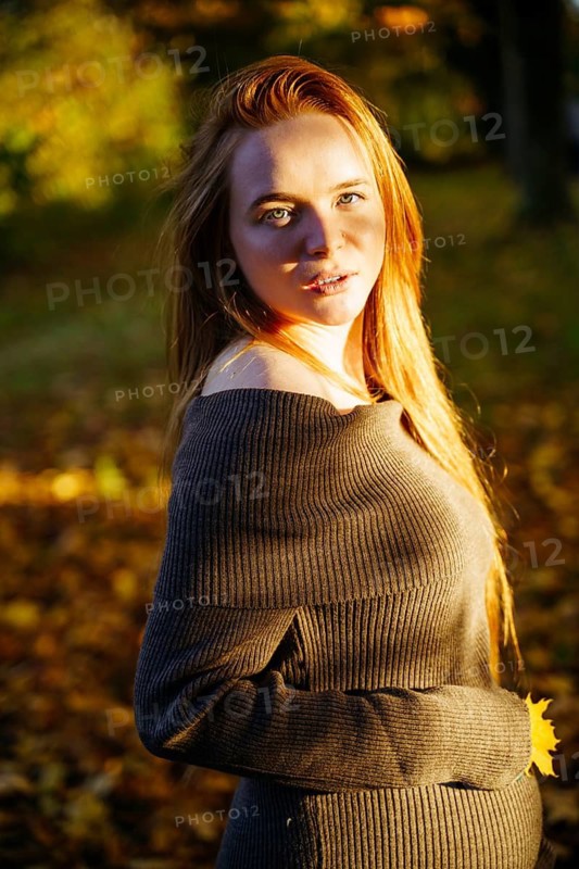 A beautiful pregnant woman with flowing red hair stands in a sunlit park, surrounded by autumn leaves. The warm sunlight highlights her serene expression and natural beauty
