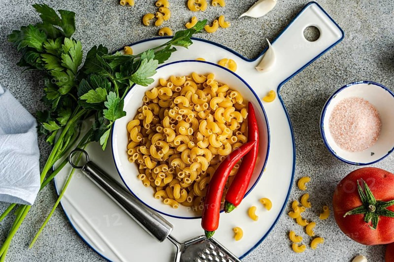 A bowl of uncooked chifferi rigati pasta is artfully displayed with fresh parsley, red chili peppers, garlic cloves, and a ripe tomato on a textured stone background