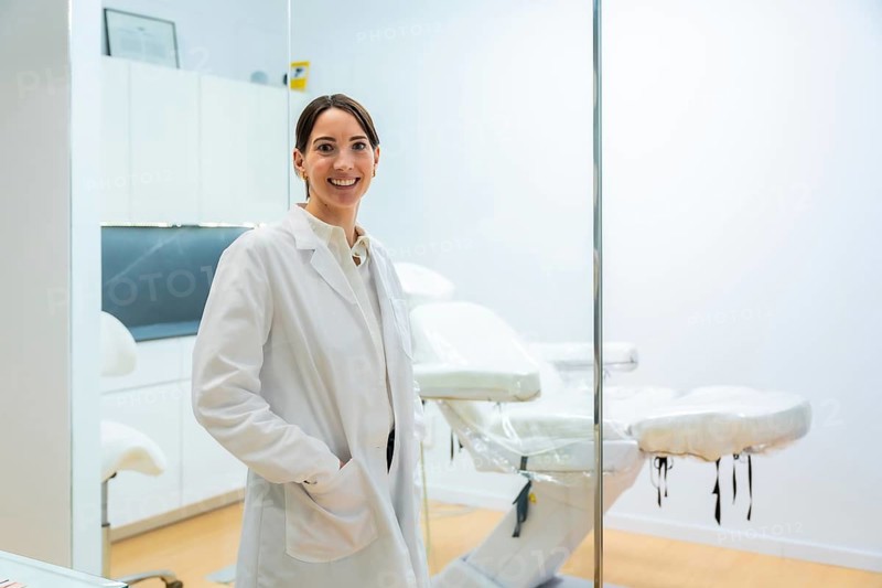 Smiling female doctor in a white coat stands in a modern aesthetic clinic, confident and professional, posing by an exam bed to represent beauty, wellness, and medical expertise