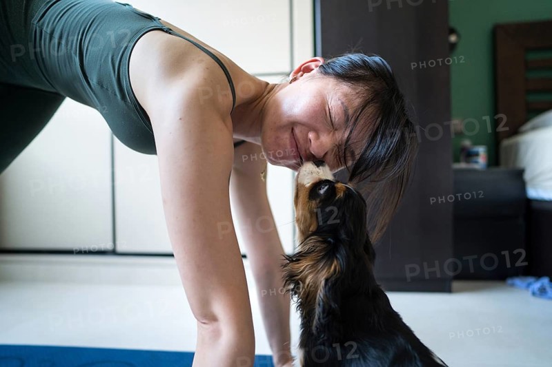 A young Asian woman attempts to exercise at home, but her playful dog interrupts her workout. This lighthearted moment captures the bond between pet and owner during a home fitness routine