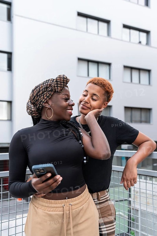 A joyful LGBT multiethnic couple shares a tender moment outdoors, leaning against a railing with smiles and affection. One holds a phone, symbolizing connection and love