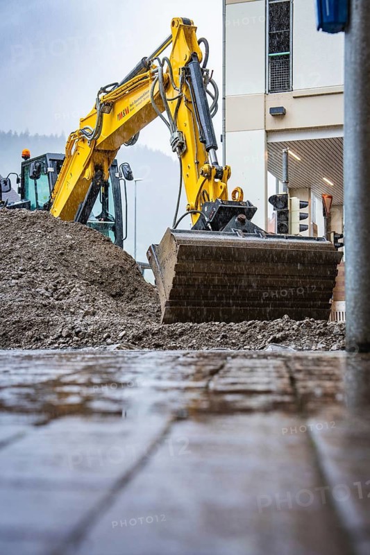 A yellow excavator doing earthworks next to a building on a wet road, Hermann Hesse railway construction site, Calw, Black Forest, Germany