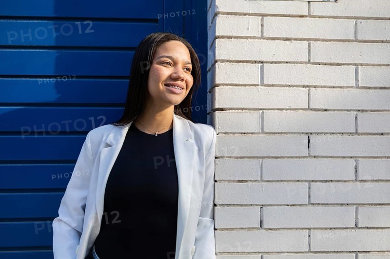Confident woman with braces in a white blazer, standing against a modern blue and white wall. Sunlit face expressing positivity and professionalism in an urban setting