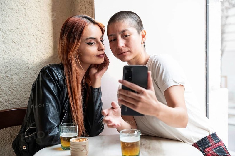 A lesbian couple enjoys drinks at a cafe, engrossed in a phone, sharing a moment of laughter and connection. They sit closely, exuding warmth and companionship