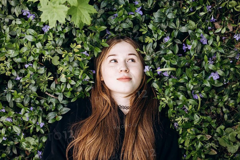 A thoughtful teenage girl lies on a lush bed of green periwinkle leaves and violet blossoms, surrounded by nature in a peaceful and reflective moment outdoors
