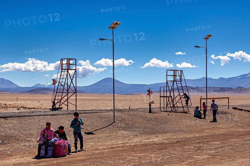 People and wooden structures can be seen in a vast desert landscape with blue skies and mountains in the background, railway station in the town of Ollagüe in the Chilean Atacama Desert