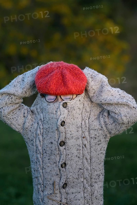 An unrecognizable young boy in a knitted sweater and red beret covers her face, standing in a park filled with autumn colors