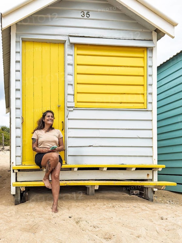 A woman enjoys a sunny day while sitting at the vibrant Brighton Bathing Boxes. These iconic structures are located on the picturesque beach in Melbourne, Australia