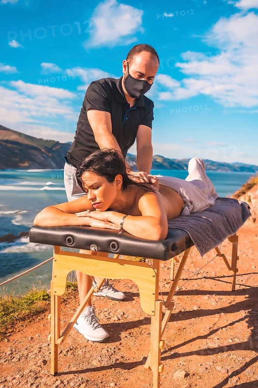 A young man performing a massage in nature on the coast near the sea, masseuse with a face mask in the coronavirus pandemic