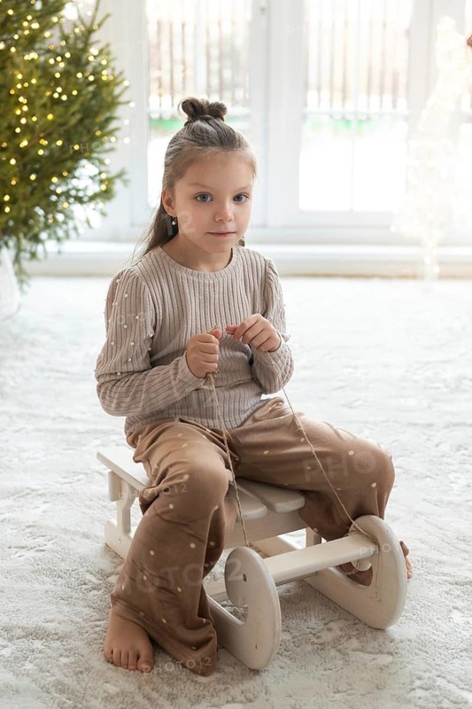A young girl sits on a wooden toy in a cozy room, decorated with a Christmas tree, during holiday celebrations. She wears a beige sweater and brown trousers, looking directly at the camera with a subtle smile
