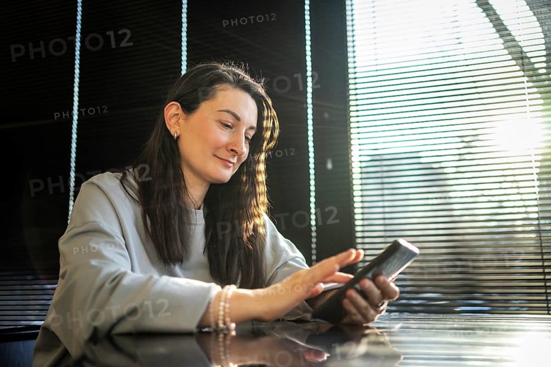 A mid-aged woman enjoys browsing on her smartphone while seated at a cafe bathed in sunlight filtering through blinds