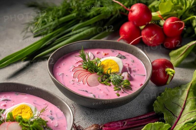 A bowl of chilled beetroot soup garnished with a slice of hard-boiled egg, fresh dill, and radish slices, surrounded by raw radishes and greens on a textured surface