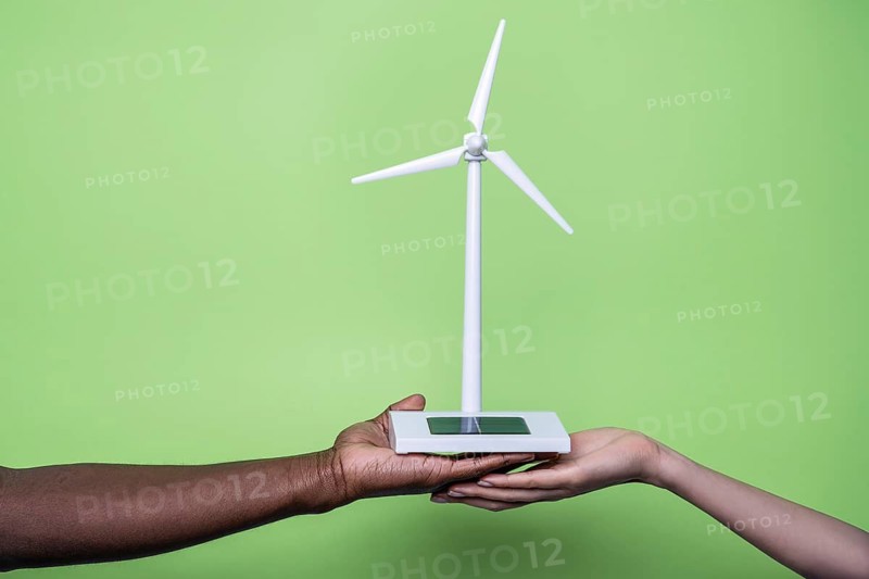 Close-up of diverse hands holding a small wind turbine model against a vibrant green background, symbolizing unity and commitment to sustainable and renewable energy practices