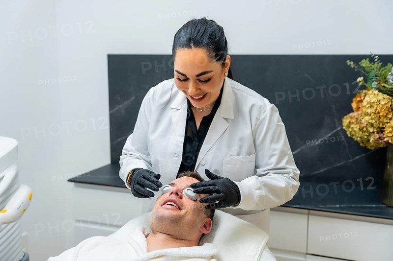 Aesthetician applying protective eyewear on a smiling man lying down during a facial treatment procedure at a modern aesthetic clinic for skin care and wellness
