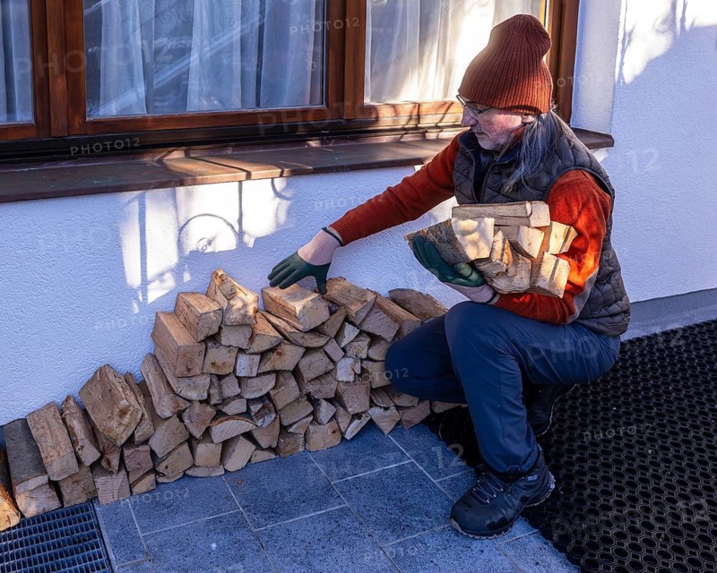 An elderly man in winter attire and gloves carefully arranges eco-friendly firewood outside his home, demonstrating sustainable domestic practices in a residential setting