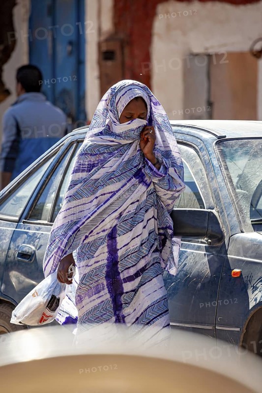A woman in a traditional dress walks past a car in an urban environment, Veiled woman at the Tamanrasett festival in the Sahara in Algeria