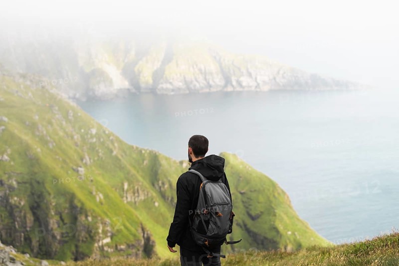 A hiker with a backpack stands on lush green cliffs overlooking the Norwegian coast Misty fog rolls over the dramatic landscape, creating a serene and breathtaking atmosphere