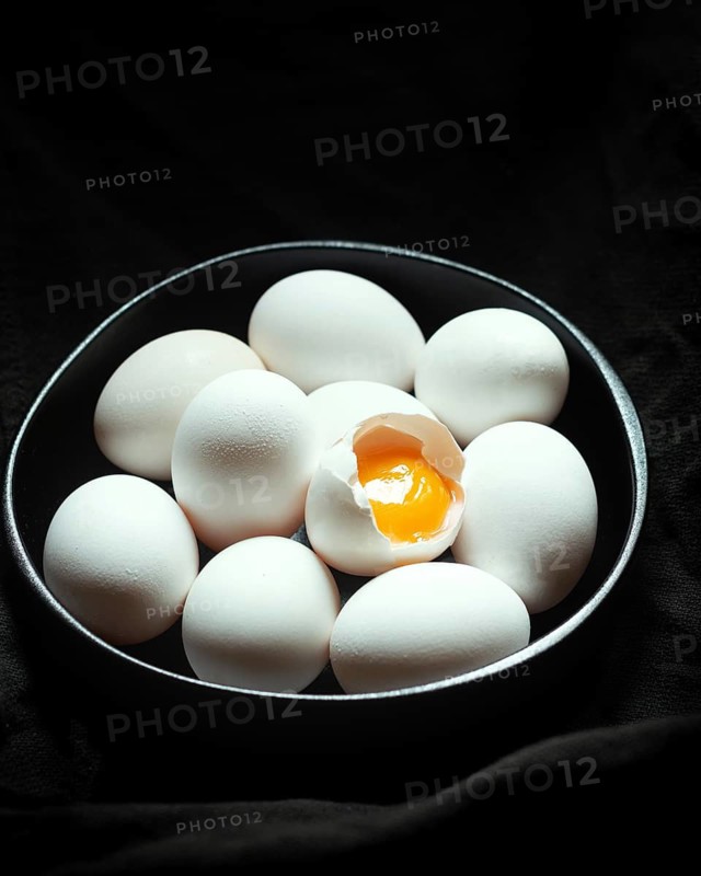 A collection of white eggs sits encircled in a dark bowl on a black background, with one egg cracked open to reveal its bright yellow yolk, adding a pop of color