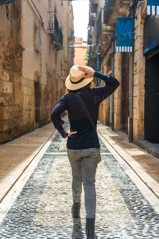 Tourist walking in the medieval city of tarragona, spain, holding her straw hat and looking up at the old buildings