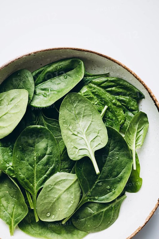 Fresh green spinach leaves with glistening dewdrops on a round, neutral-toned ceramic plate, providing a refreshing and healthy food theme in natural lighting