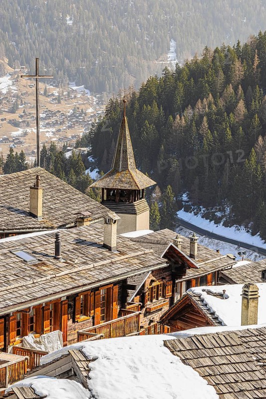 Snow-covered roofs of old farmhouses and barn storage, in the back the large wooden cross and the church tower of the Saint Theodule church, Grimentz, Val d'Anniviers, Valais Alps, Canton of Valais, Switzerland
