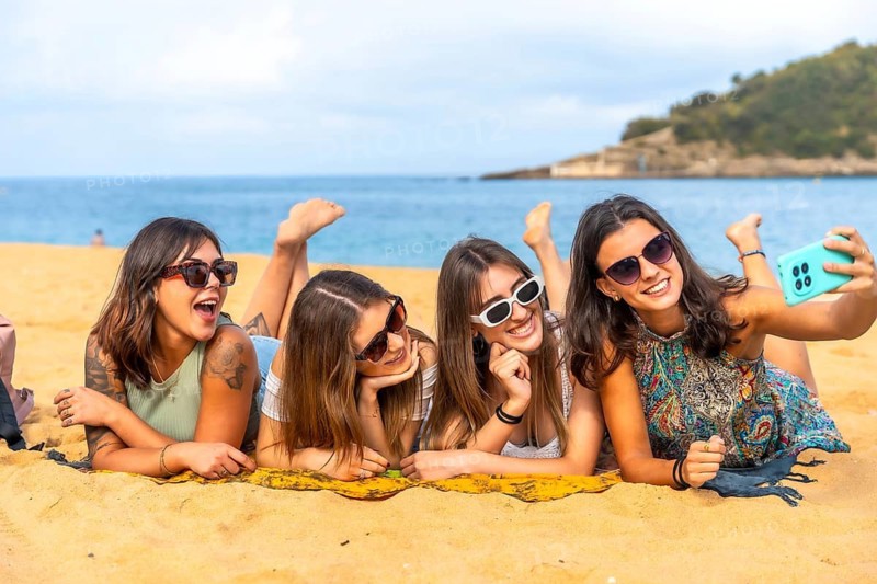 Group of young women lying on warm sand, smiling and taking a selfie with a smartphone, enjoying sunny beach vacation, laughing, relaxing and capturing joyful friendship moments