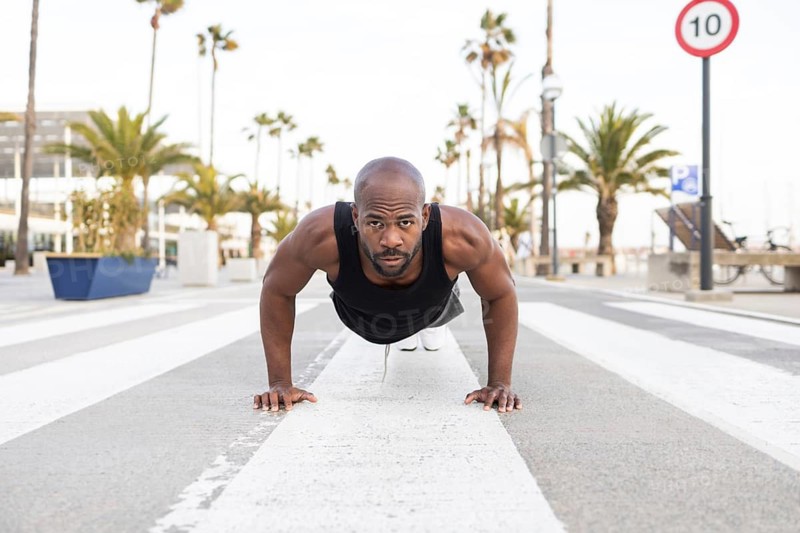 A fit cuban man performs push ups on a city street, surrounded by palm trees and urban scenery. He is focused on maintaining strength and fitness in an outdoor setting