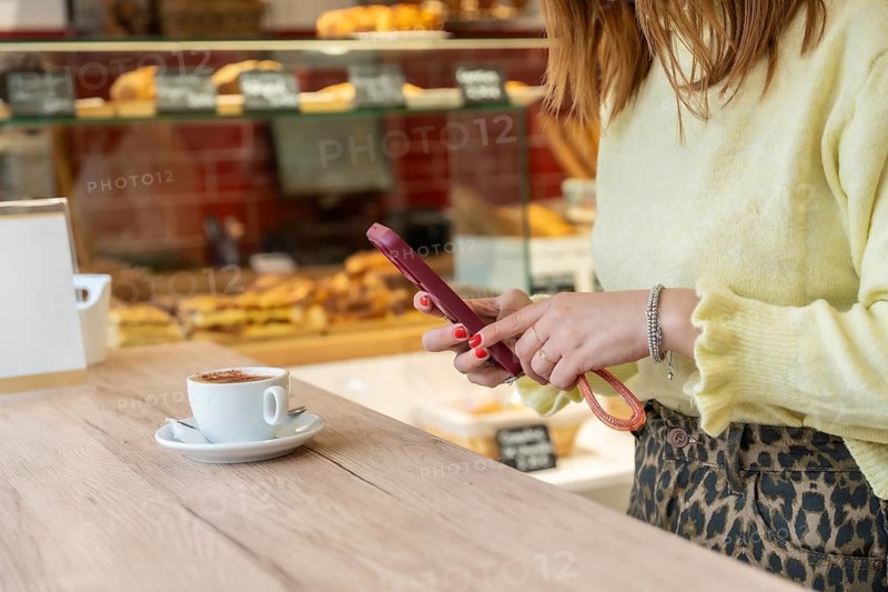 Woman standing at a bakery counter, looking at her smartphone while enjoying a cup of coffee, showcasing modern connectivity and leisure in a relaxed setting