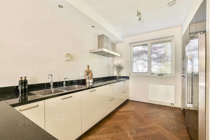A sleek, modern kitchen showcasing minimalist design. Features include a black counter top, dual sinks, wooden herringbone floor, and stainless steel appliances under natural light