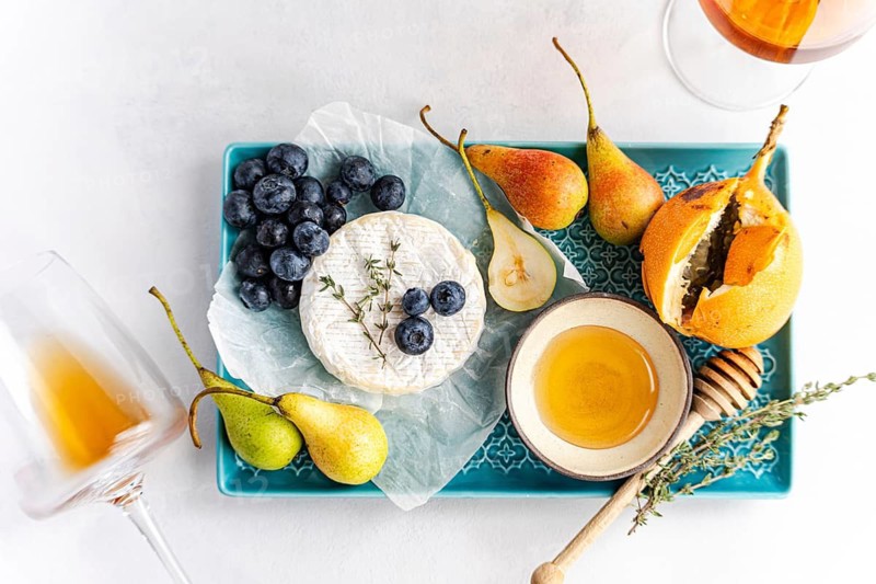 A beautifully arranged gourmet platter featuring creamy brie cheese, fresh blueberries, sliced pears, and a drizzle of honey on a decorative tray, accompanied by a glass of amber wine