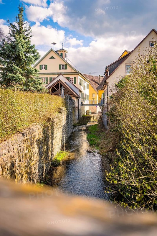 Small river flows between historic houses surrounded by green bushes under cloudy sky, Deufringen, Aidlingen, Böblingen district, Germany