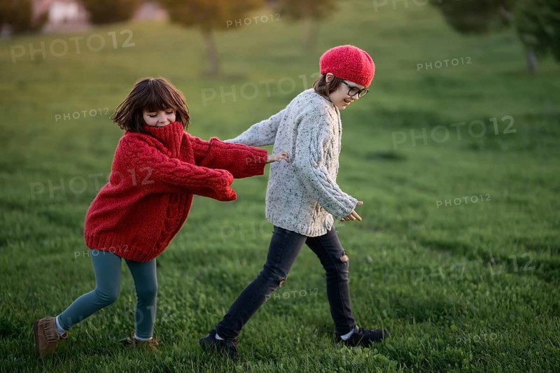 Two siblings, a girl in a red sweater and a boy in a gray sweater, enjoy a playful day in a park during autumn, highlighting the joy and warmth of the season