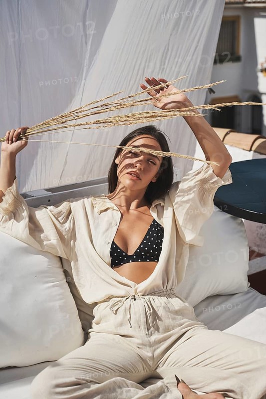 A woman enjoys a sunny day on a stylish rooftop, dressed in casual summer attire She relaxes on comfortable cushions, holding wheat stalks, embodying a chic, carefree lifestyle