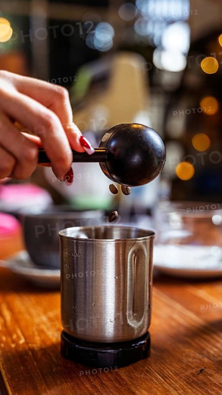 A barista scoops aromatic coffee beans into a metal cup in a warm, ambient cafe. The scene captures the essence of handcrafted coffee preparation in a cozy cafe environment
