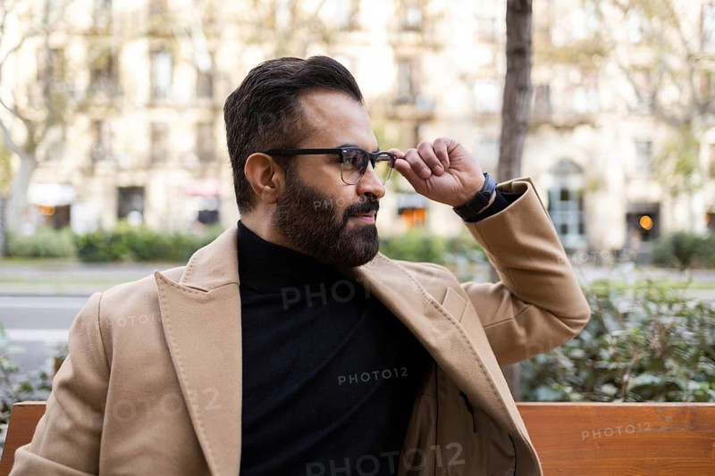 Stylish Indian man wearing glasses, a turtleneck, and a beige coat adjusts his glasses while seated on a bench in a city during winter