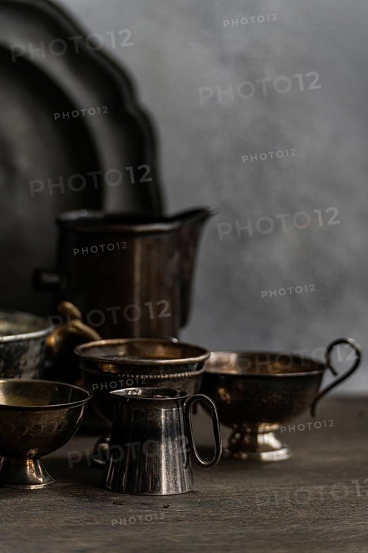 A collection of vintage metal cups and dishes arranged on a textured surface against a muted background. The worn metallic surfaces reveal their rustic charm and elegance