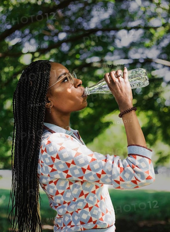 A woman enjoys a sunny day in nature, hydrating with water and capturing the moment with her camera. It's a perfect summer day for outdoor exploration and creativity