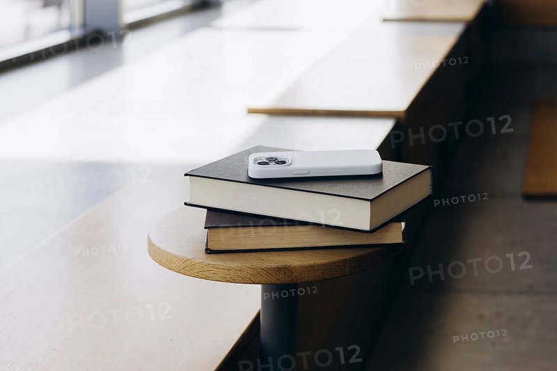 A modern smartphone rests on a stack of two hardcover books placed on a wooden bench. The setting appears to be an indoor space, with natural light streaming in through windows