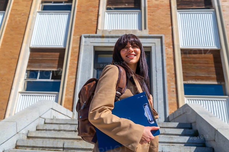 Young female student smiling, carrying a backpack and clipboard, standing on stone stairs in front of a university building, representing modern education and academic life