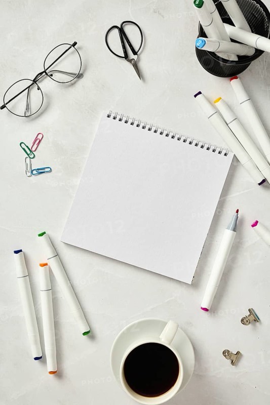 Top view of a sketchbook accompanied by colorful markers, glasses, scissors, paper clips, and a coffee cup on a textured marble table, depicting a creative workspace setup