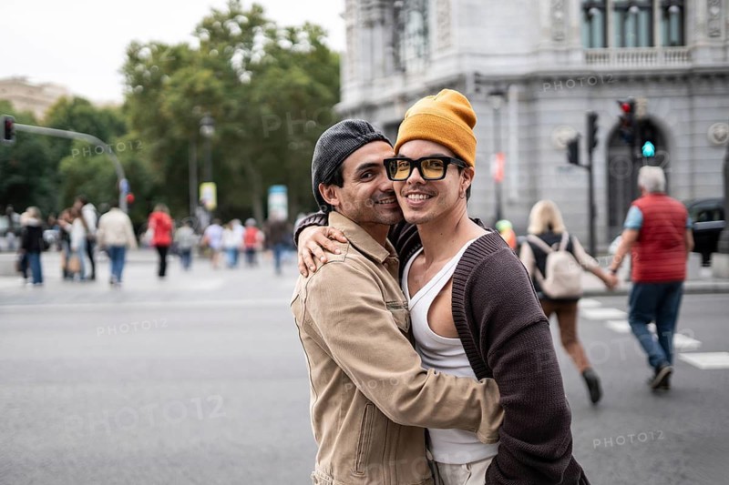 A joyful Colombian gay couple embraces warmly on a bustling city street. They smile brightly, showcasing love and connection in an urban setting with people in the background