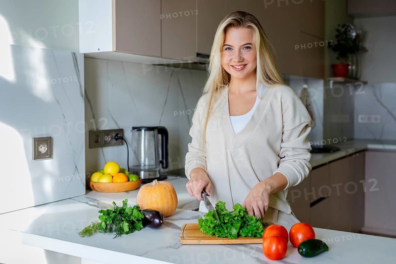 A woman with a friendly smile is chopping fresh lettuce on a wooden board in a modern kitchen. Surrounding her are a variety of colorful vegetables and fruits healthy