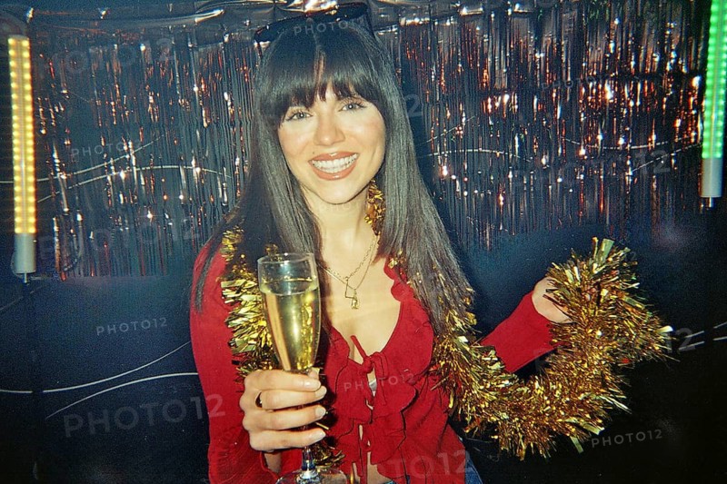 Happy young woman smiling and holding a champagne flute with golden tinsel, celebrating a festive holiday party indoors with shimmering decorations and vintage film aesthetic
