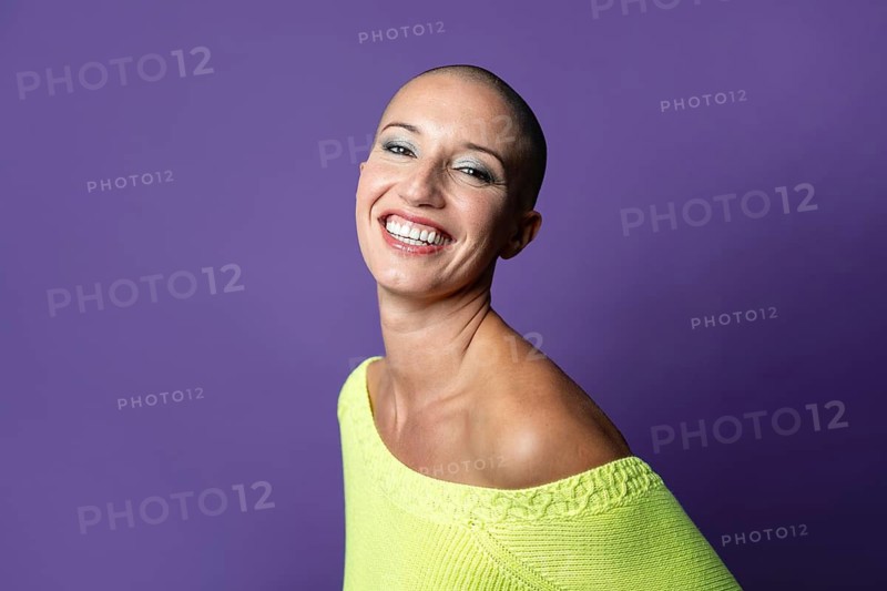 A middle-aged woman with a shaved head smiles confidently against a vibrant purple background, wearing a bright yellow top. Her expression radiates positivity and self-assurance