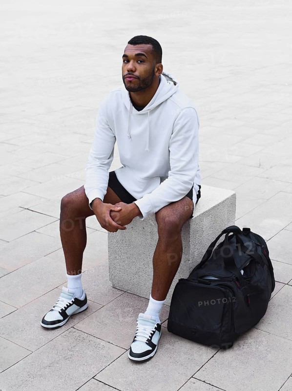 A young black male athlete sitting on a concrete block outdoors. He wears a white hoodie and sneakers, with a sports bag beside him. Relaxed, poised expression