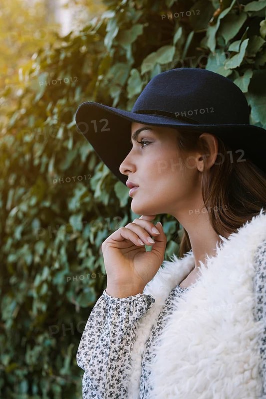 A woman in a black hat, floral-patterned blouse, and white fur vest, gazes thoughtfully against a lush green leafy backdrop, capturing a serene outdoor moment