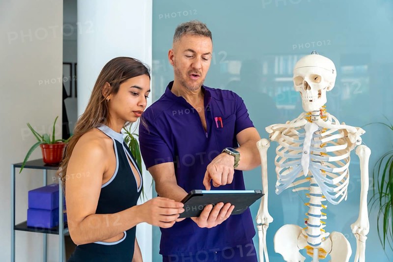 Physiotherapist consulting female patient about injury diagnosis and rehabilitation plan, using a tablet and human skeleton model to illustrate medical recovery concepts