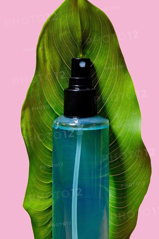 A striking blue cosmetic spray bottle nestled between the curves of a vivid green leaf, highlighted by a soft pink background. Ideal for beauty and skincare products