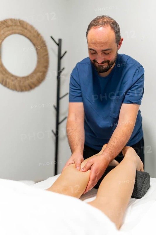 Male masseur performing leg massage on a client lying on a massage table in a bright treatment room, emphasizing muscle relief, physical therapy and relaxation benefits