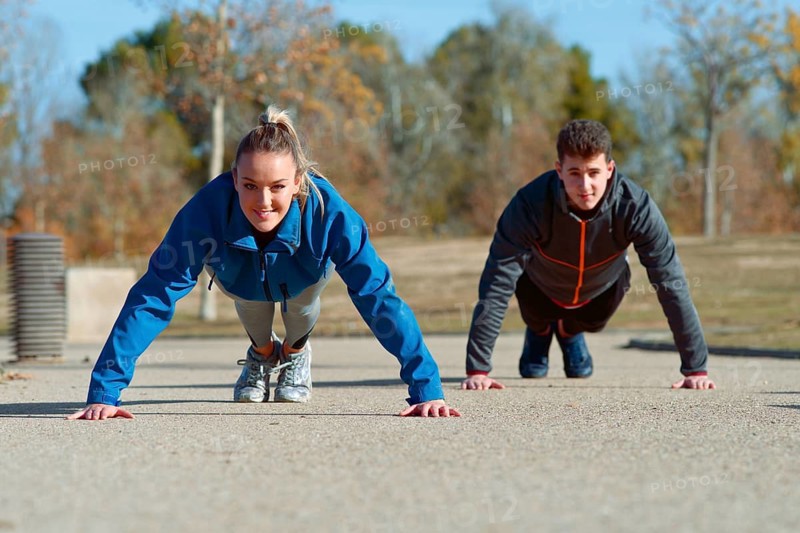 A man and woman exercises outdoors on a sunny day, performing push ups on a concrete path in the park. They wear athletic gear, embodying a fitness focused lifestyle